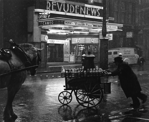 Black-and-white photo of a rainy street scene with a man pushing a cart while a horse stands nearby, in front of a theater marquee displaying movie titles.