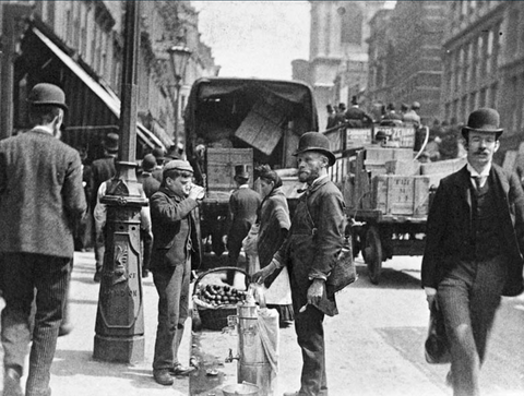 Early 20th-century street scene with children working and adults walking, including a street vendor and vintage vehicles.