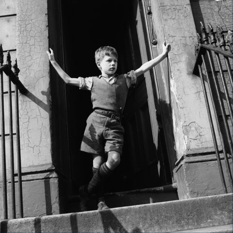 Young boy in vintage clothing standing at the top of steps with his arms stretched wide, in a doorway of an old building.