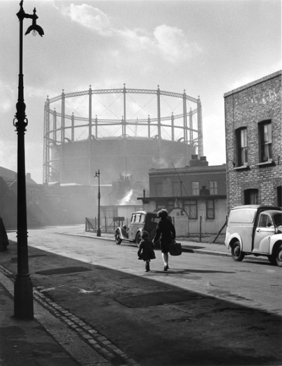 Black and white photo of a city street with two people walking, gas holder in the background, and vintage cars parked.