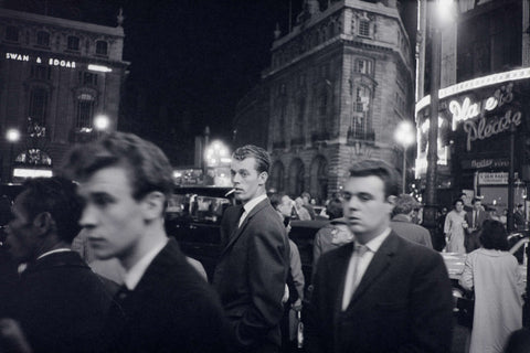 Black and white photo of a bustling city street at night with people walking, neon signs lit, and a man in the center looking back towards the camera.