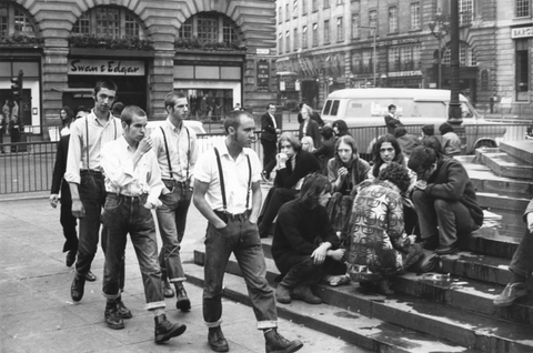 Young people, some in punk fashion, gather and socialize on a city street, buildings and a van visible in the background.