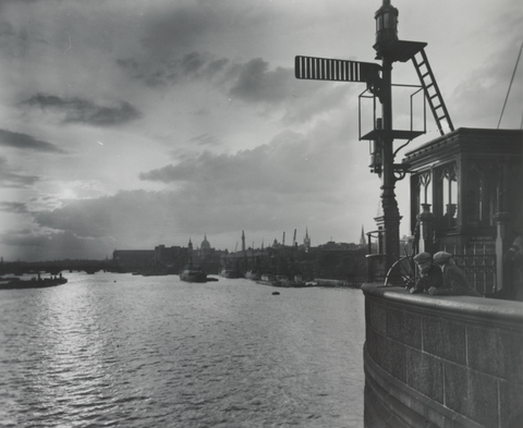 Black and white photo of a river view from a bridge, featuring distant city skyline and a bicycle leaning against railing under a lamp post.