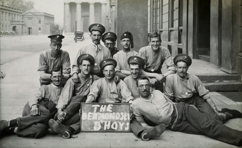 A group of nine early 20th-century firefighters in uniform, sitting and lying together, holding a sign that reads "The Bertholdsen Boys.