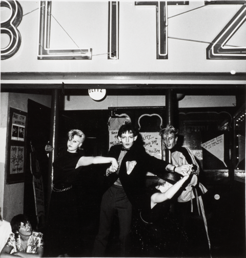 A group of four people posing dramatically under a sign reading "BLITZ" at a nightclub entrance, with one person sitting on the ground in front of them.