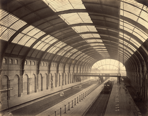 Vintage sepia-toned photograph of an empty train station with arched glass roof and multiple tracks, taken in the early 20th century.