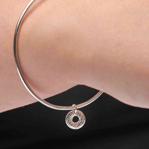 Person wearing a silver bangle with a circular charm on their wrist against a black background