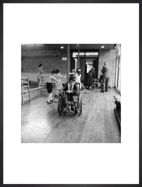 A disabled young boy using a wheelchair rolls a hoop along the floor during a physical education lesson at the 'FDR School for Handicapped Children', NW3