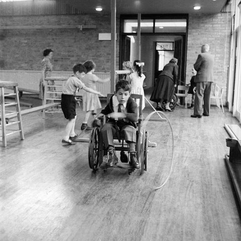 A disabled young boy using a wheelchair rolls a hoop along the floor during a physical education lesson at the 'FDR School for Handicapped Children', NW3