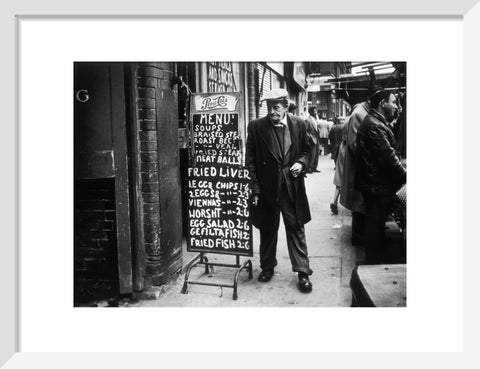 A man reads a chalk board on the street outside a cafe1961