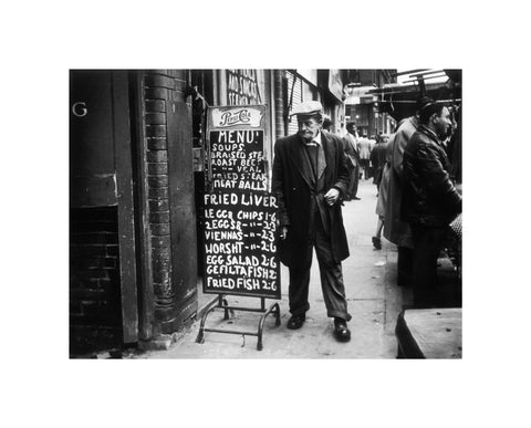 A man reads a chalk board on the street outside a cafe1961