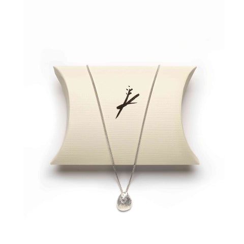 Silver necklace with an irregular round pendant resting against a beige pillow pouch with a stylised black logo 