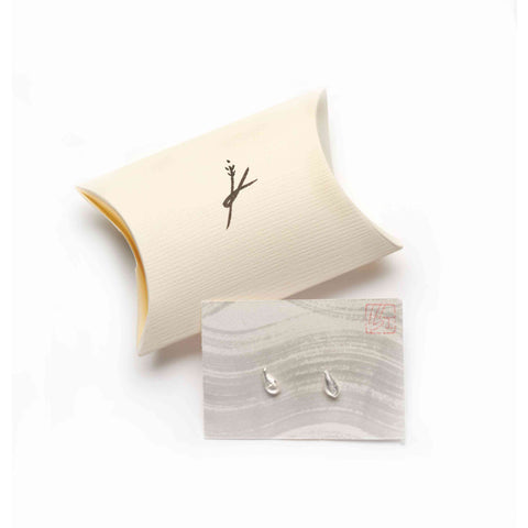Pair of silver teardrop-shaped stud earrings on a grey wavy-patterned card with visible red logo leaning against beige pillow pouch on a white background