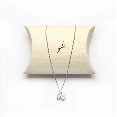 Silver necklace with two irregular teardrop-shaped pendants on a beige pillow pouch with a black stylised logo 