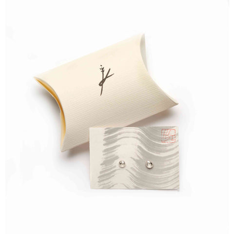 Pair of irregular silver stud earrings  on a wavy patterned card with a visible brand logo leaned against a beige pillow pouch with a black stylised logo.