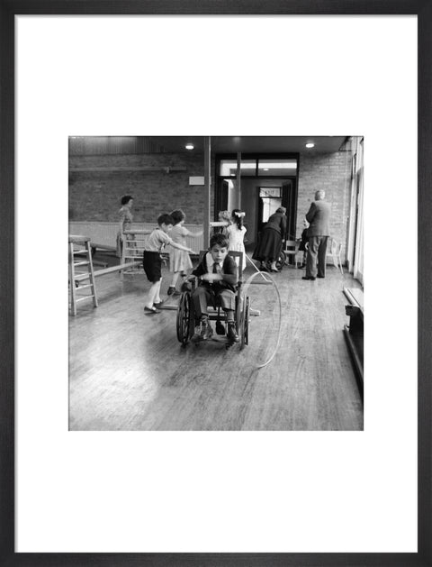 A disabled young boy using a wheelchair rolls a hoop along the floor during a physical education lesson at the 'FDR School for Handicapped Children', NW3