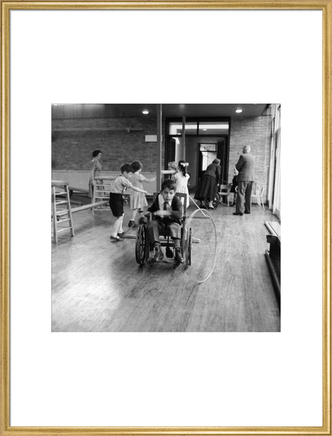 A disabled young boy using a wheelchair rolls a hoop along the floor during a physical education lesson at the 'FDR School for Handicapped Children', NW3