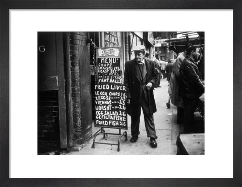 A man reads a chalk board on the street outside a cafe1961