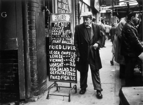 A man reads a chalk board on the street outside a cafe1961