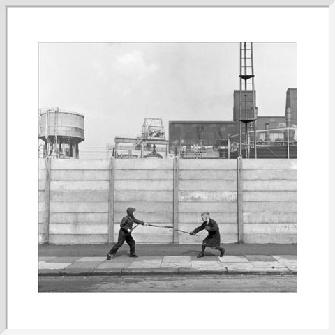 Two boys fighting with sticks in front of a concrete wall. c.1955